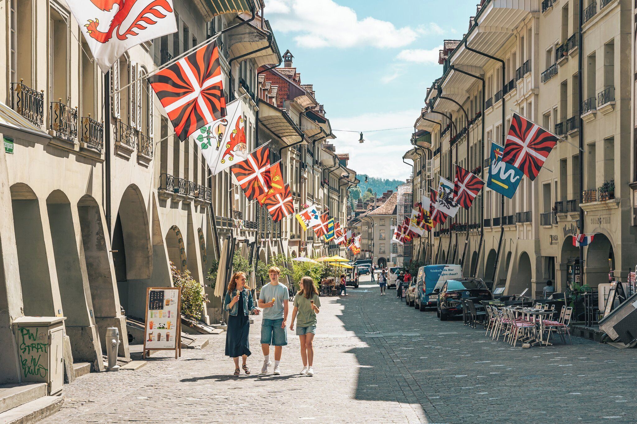 A cobblestone street lined with buildings, some with balconies and arches. Flags are hanging from the buildings, and there are tables and chairs set up on the sidewalk, suggesting a public space or café.
