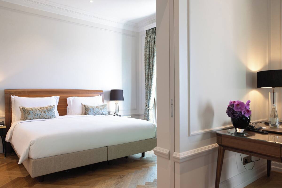 A hotel room with a large bed, two bedside tables with lamps, a small desk with a lamp, and a vase with flowers.
