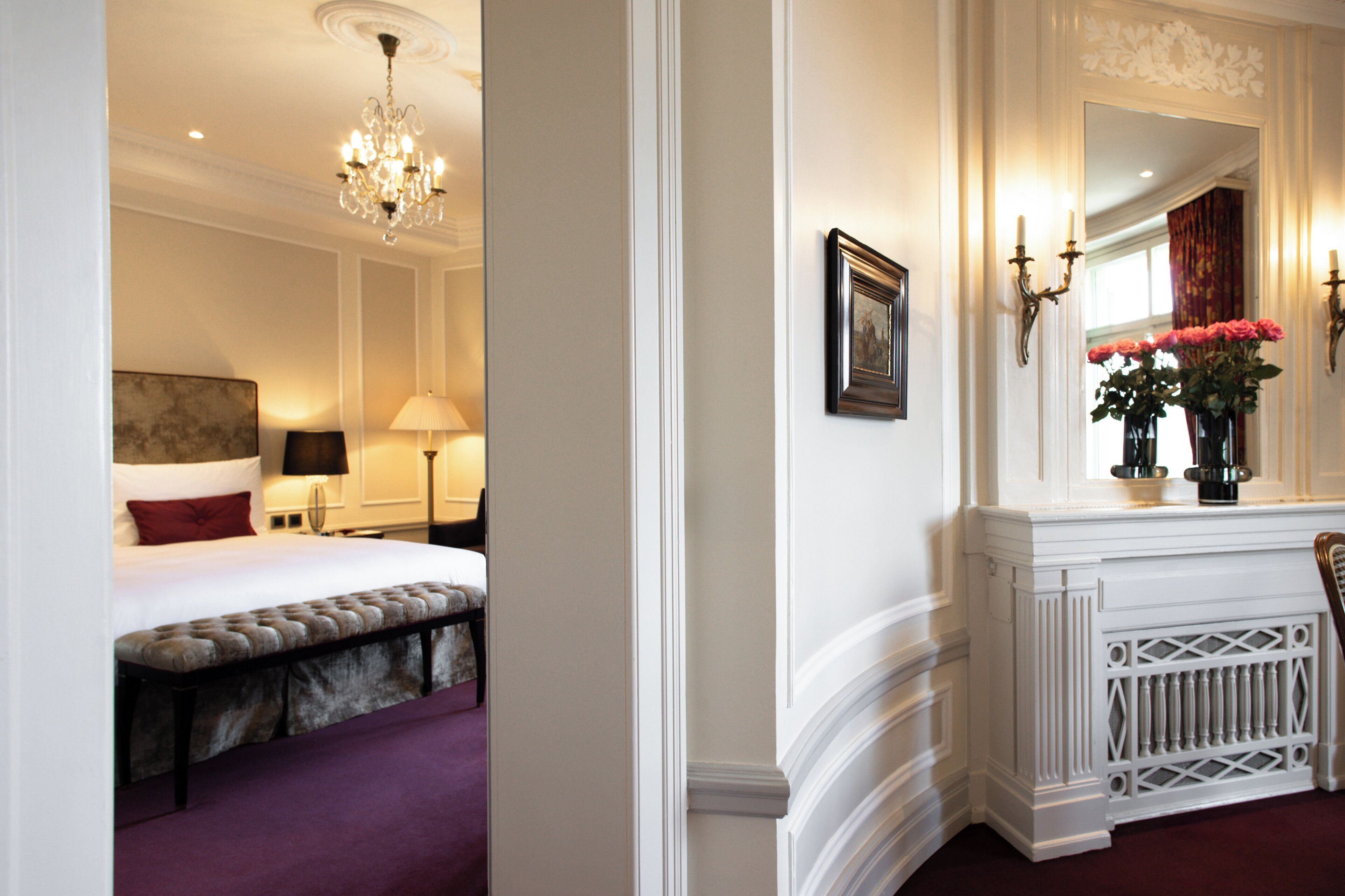 A hotel room with a bed, bench, chandelier, lamp, framed picture, mirror, and a vase with flowers.