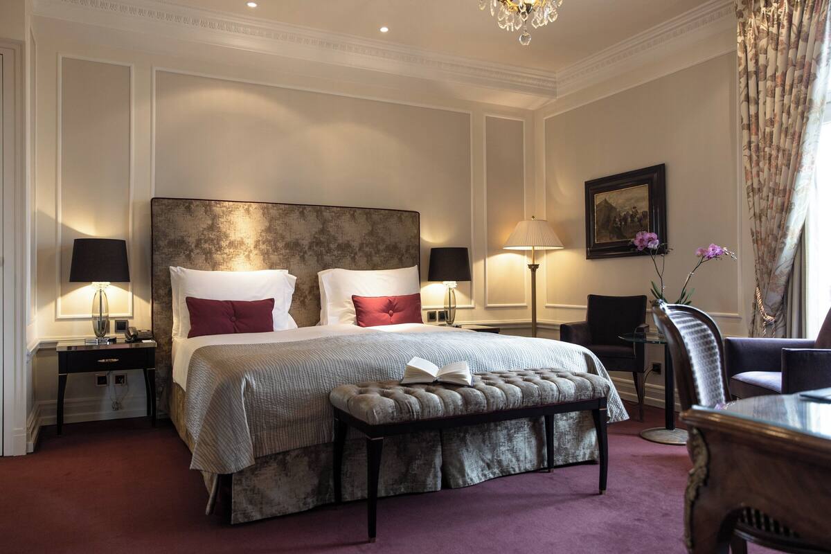A hotel bedroom with a large bed, two bedside tables, a reading chair, a small table, and a chandelier.