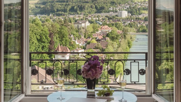 A room with a large window offering a view of a town and hills. There's a table with a vase of flowers and a glass of wine.