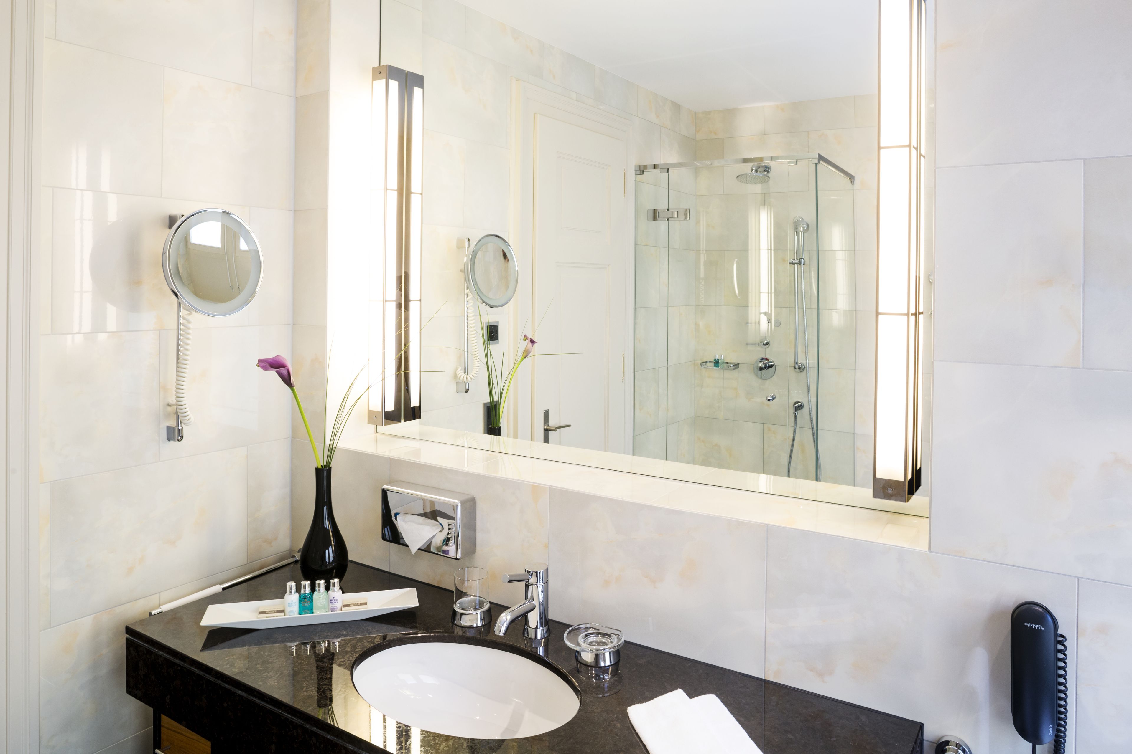 Une salle de bain moderne avec un grand miroir, un meuble-lavabo rond, une douche en verre et un téléphone.