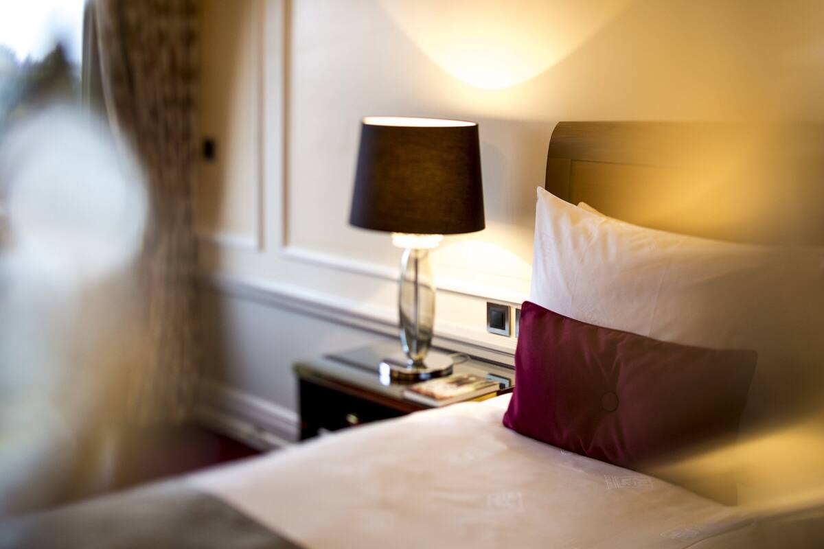 A neatly made bed with white and maroon pillows, a bedside lamp, and a nightstand with a small table.