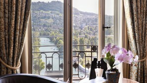 Una habitación con vistas a un río y colinas, un balcón con barandilla metálica y una orquídea en maceta sobre una mesa.