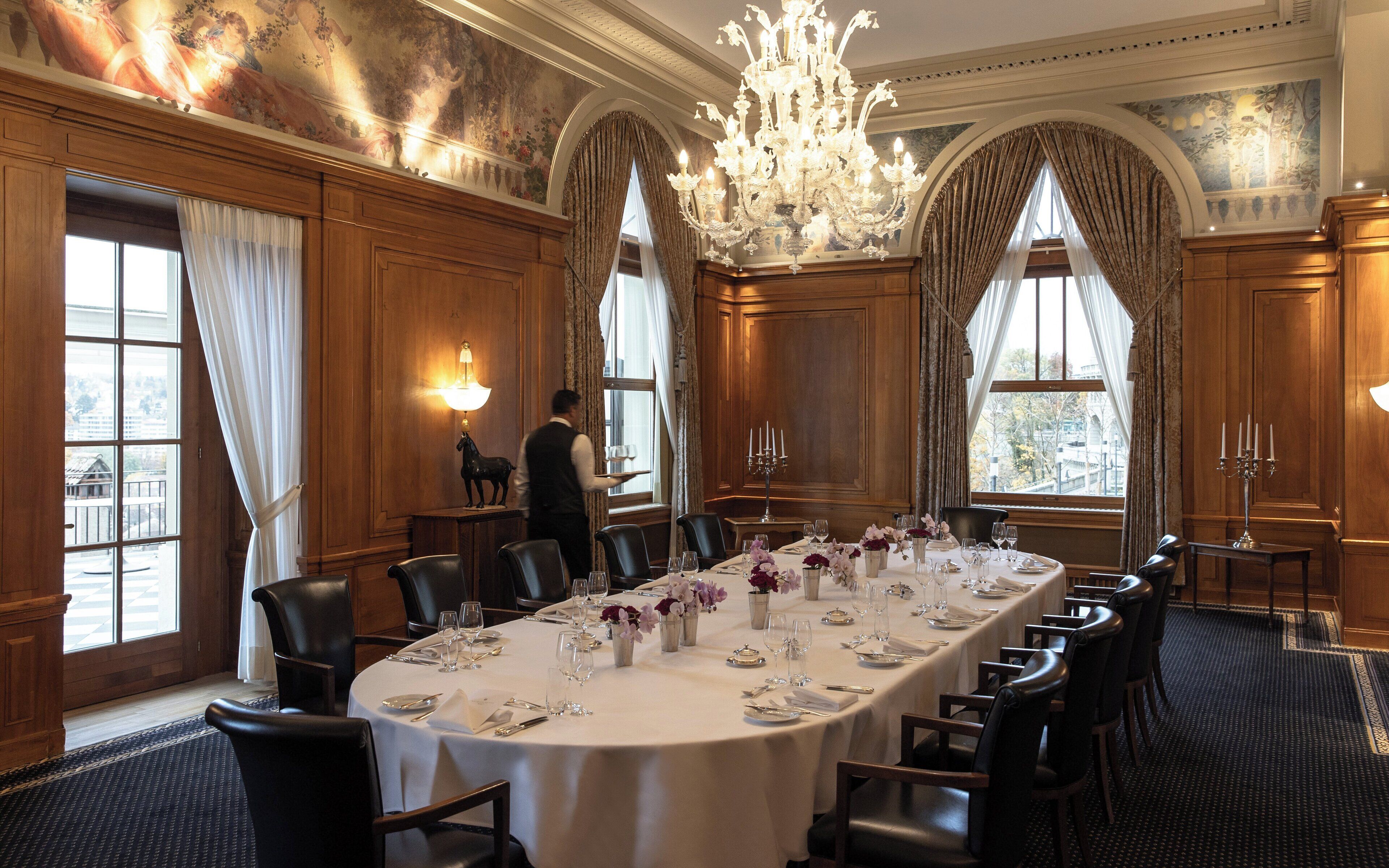 A formal dining room with a long table set for a meal, elegant chandeliers, and large windows with curtains.