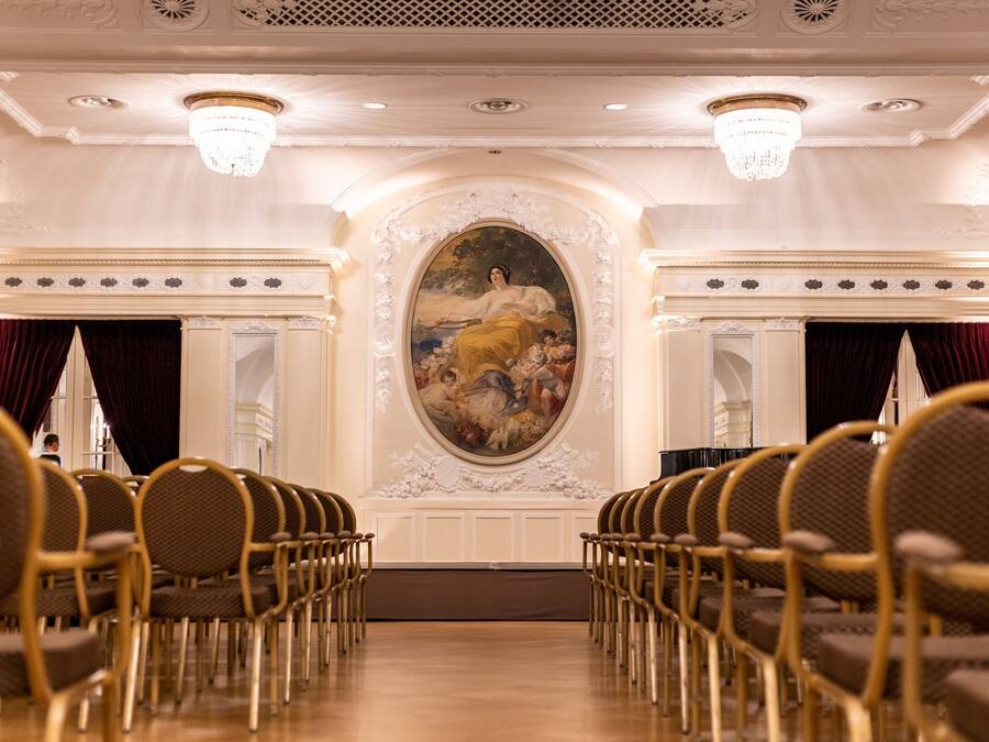 A formal room with rows of chairs, a large painting, and ornate ceiling details.