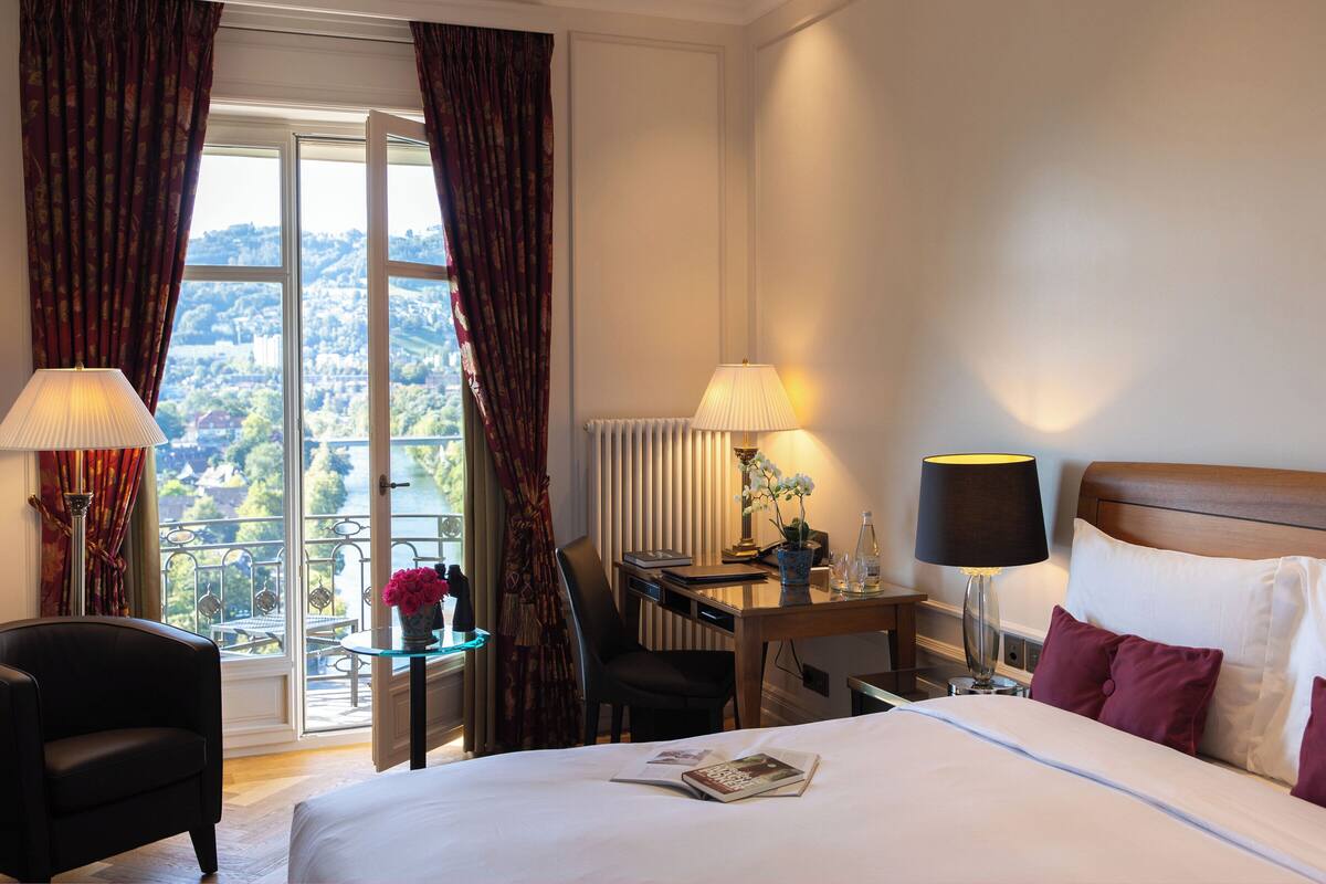 A hotel room with a bed, two lamps, a table with a vase of flowers, a chair, and a view of a river and mountains through a window.