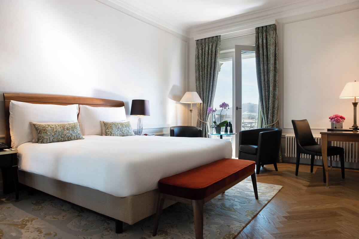 A hotel room with a large bed, a bench, a desk with a lamp, a chair, and a view of the outdoors.
