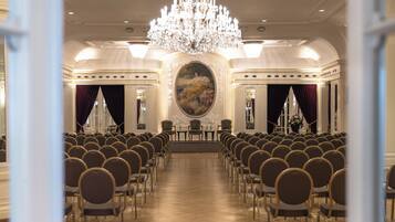 A formal room with rows of chairs, a large chandelier, and a central painting.