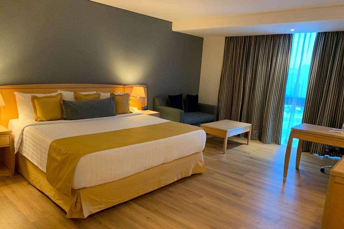 A hotel room with a large bed, a small table, a sofa, and a view of the outdoors through curtains.