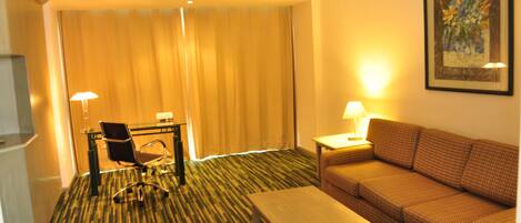 Habitación de hotel con alfombra estampada, sofá, mesita, silla, escritorio, lámpara y un cuadro en la pared.