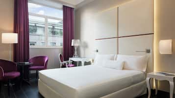 Quarto de hotel com uma cama grande, uma escrivaninha, duas cadeiras e uma janela com cortinas.