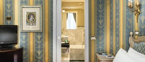 A hotel room with a bed, a television, a framed picture, a bathroom with a bathtub, and a wall-mounted light fixture.