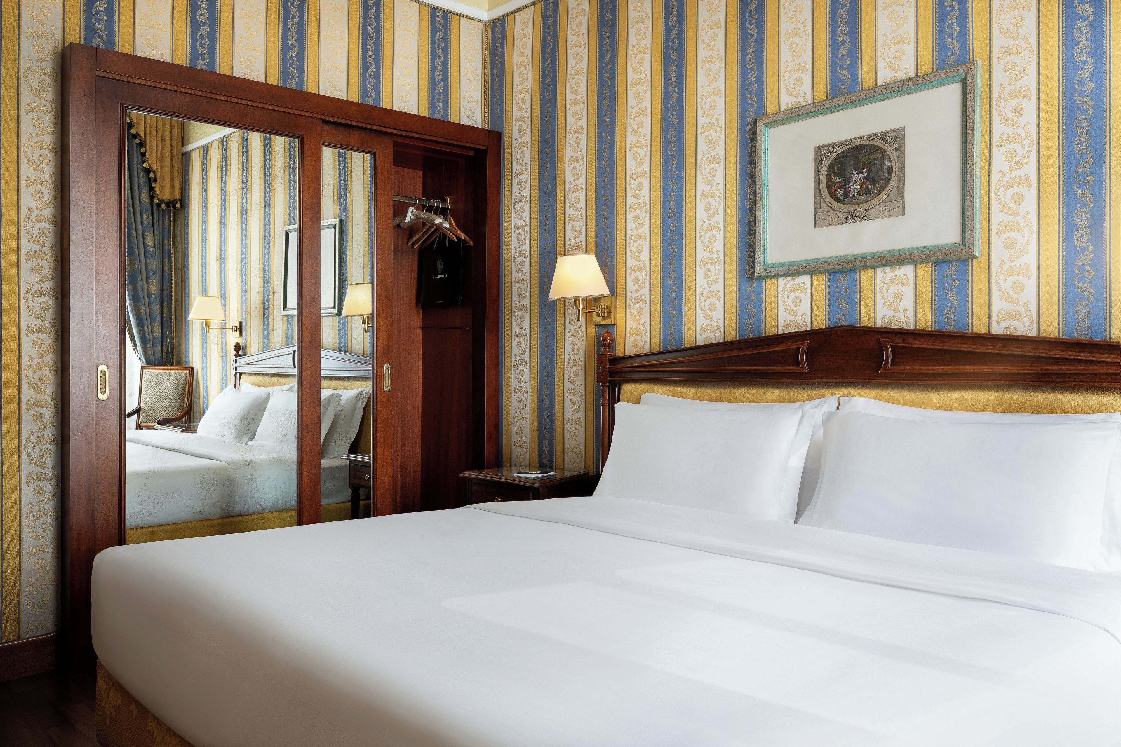 Habitación de hotel con una cama grande, cabecero de madera, espejo, un cuadro en la pared y una puerta con espejo y ropa colgada.