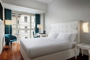 Habitación de hotel con una cama grande, una mesa pequeña, una silla y vistas a edificios a través de una ventana.