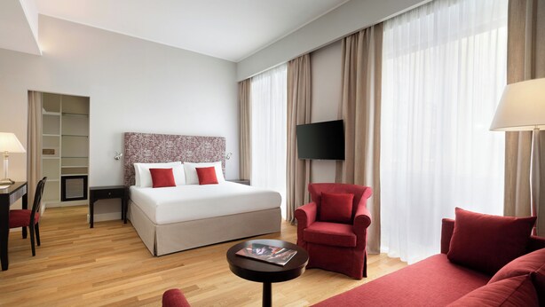 Une chambre d’hôtel avec un lit, un fauteuil rouge, un canapé, une télévision, un bureau avec une chaise et une petite table.