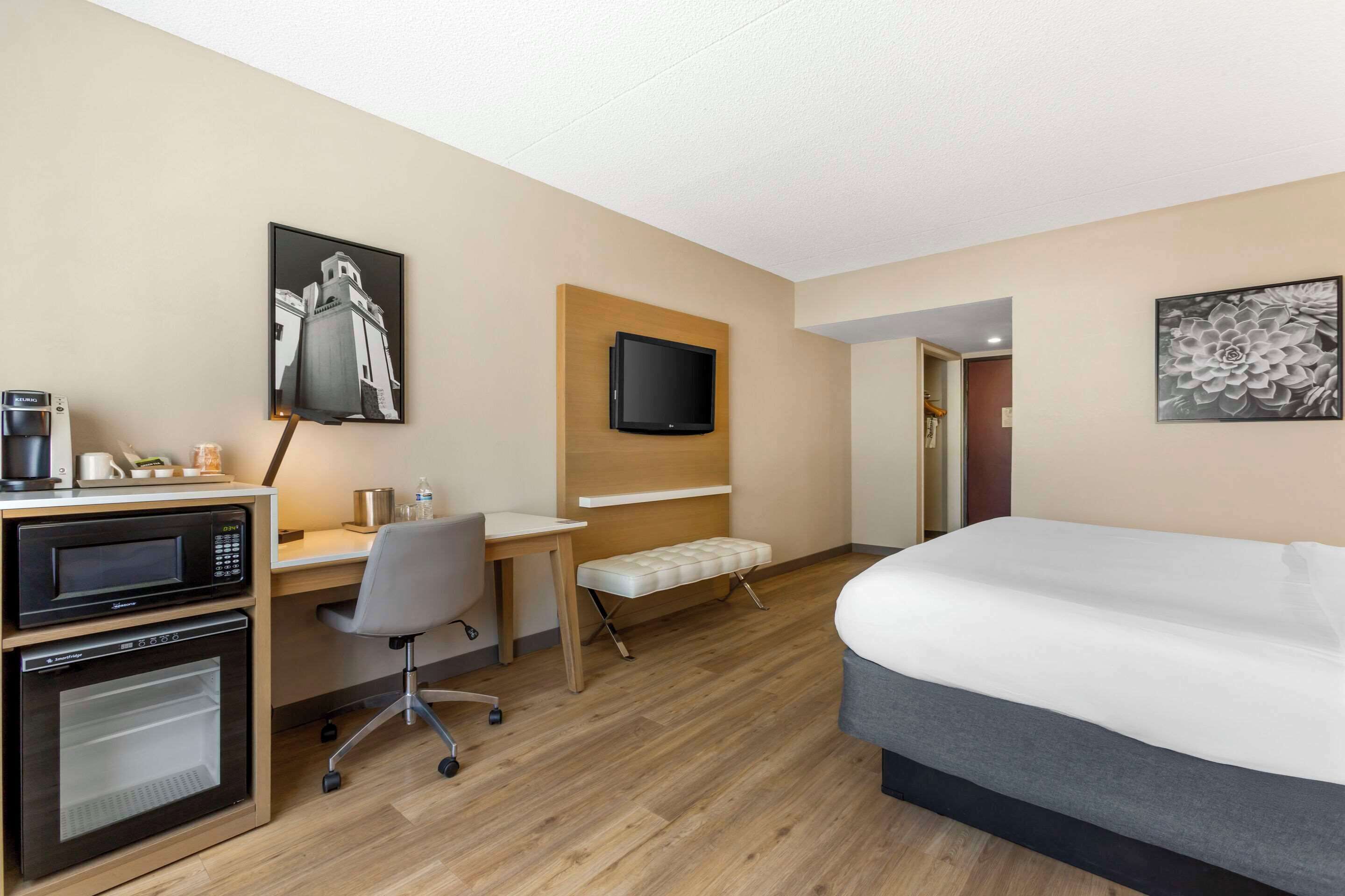 A hotel room with a bed, a desk with a microwave, a television, and a bench.