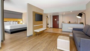 A modern hotel room with a bed, a bench, a sofa, a small table, a TV, and a kitchenette.