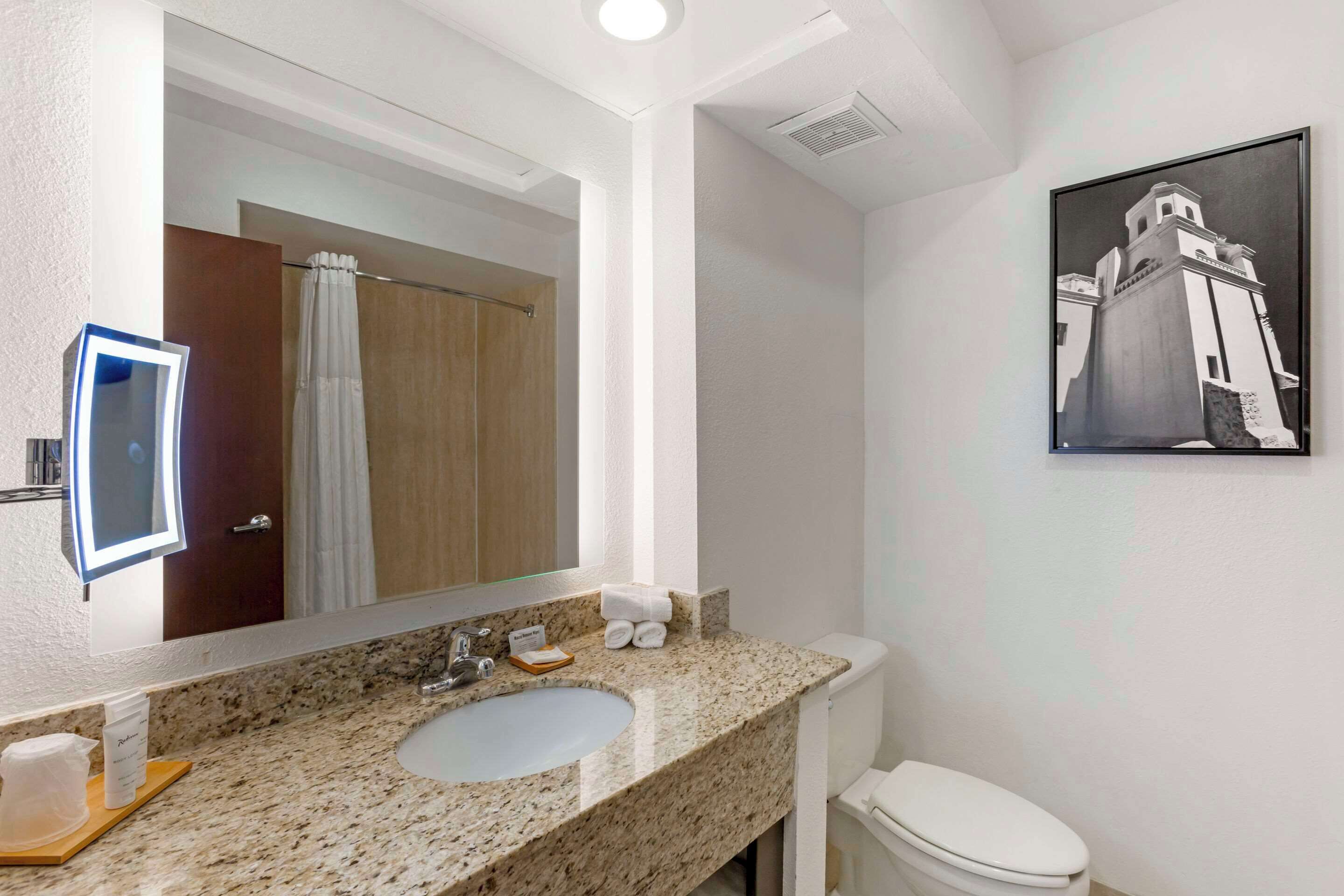 A bathroom with a granite countertop sink, a white toilet, a large mirror, and a framed picture on the wall.