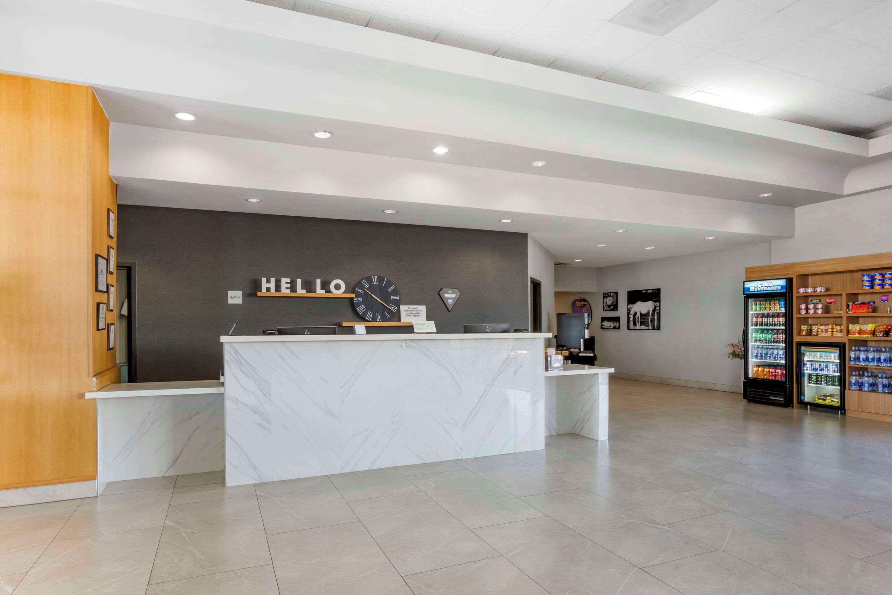 Et moderne receptionområde med en marmordisk, et ur og et skilt med teksten 'HELLO'. Der er automater og en træhylde med snacks.