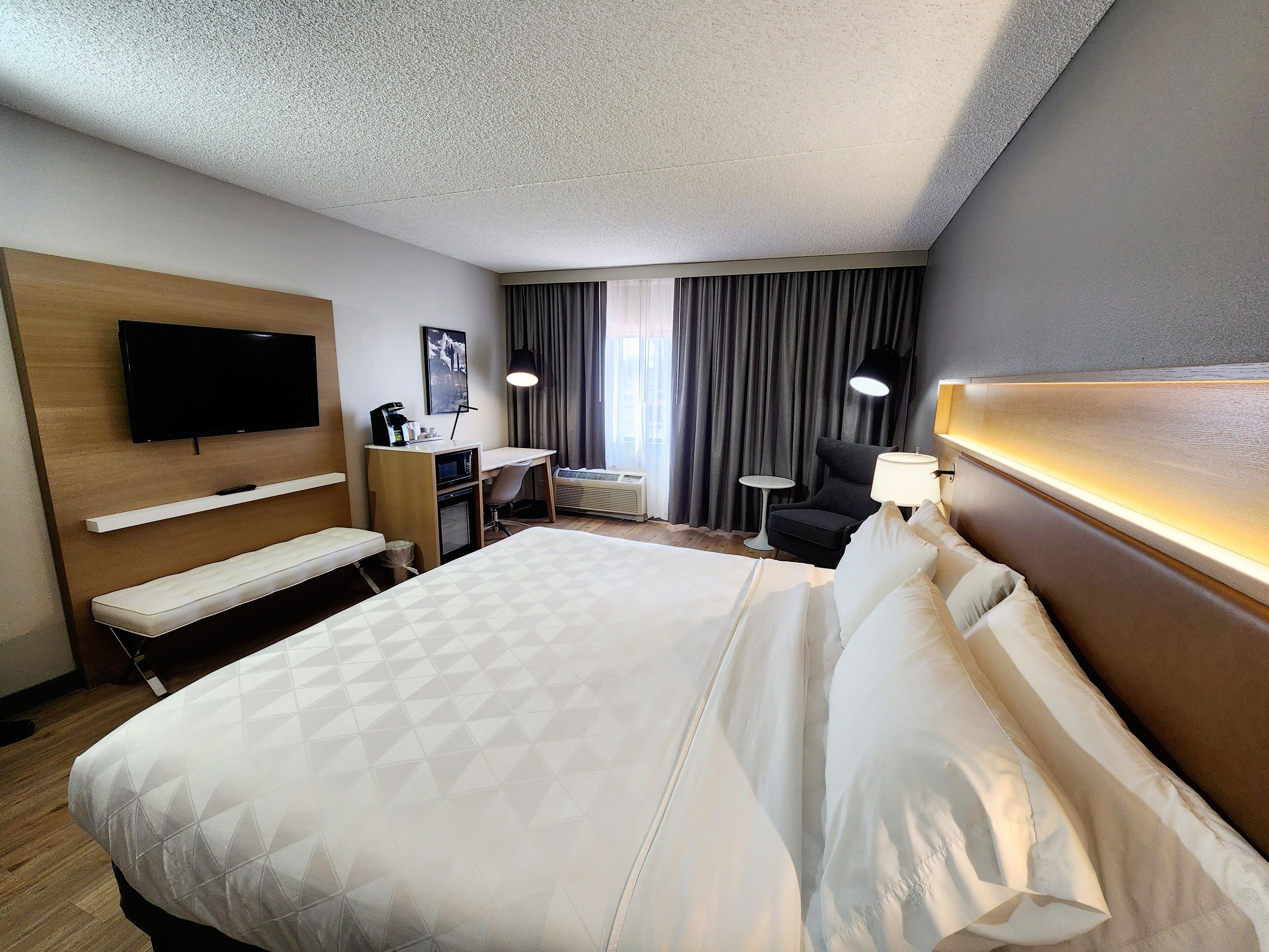 A hotel room with a large bed, a desk, a chair, a TV, a small table, and a lamp.