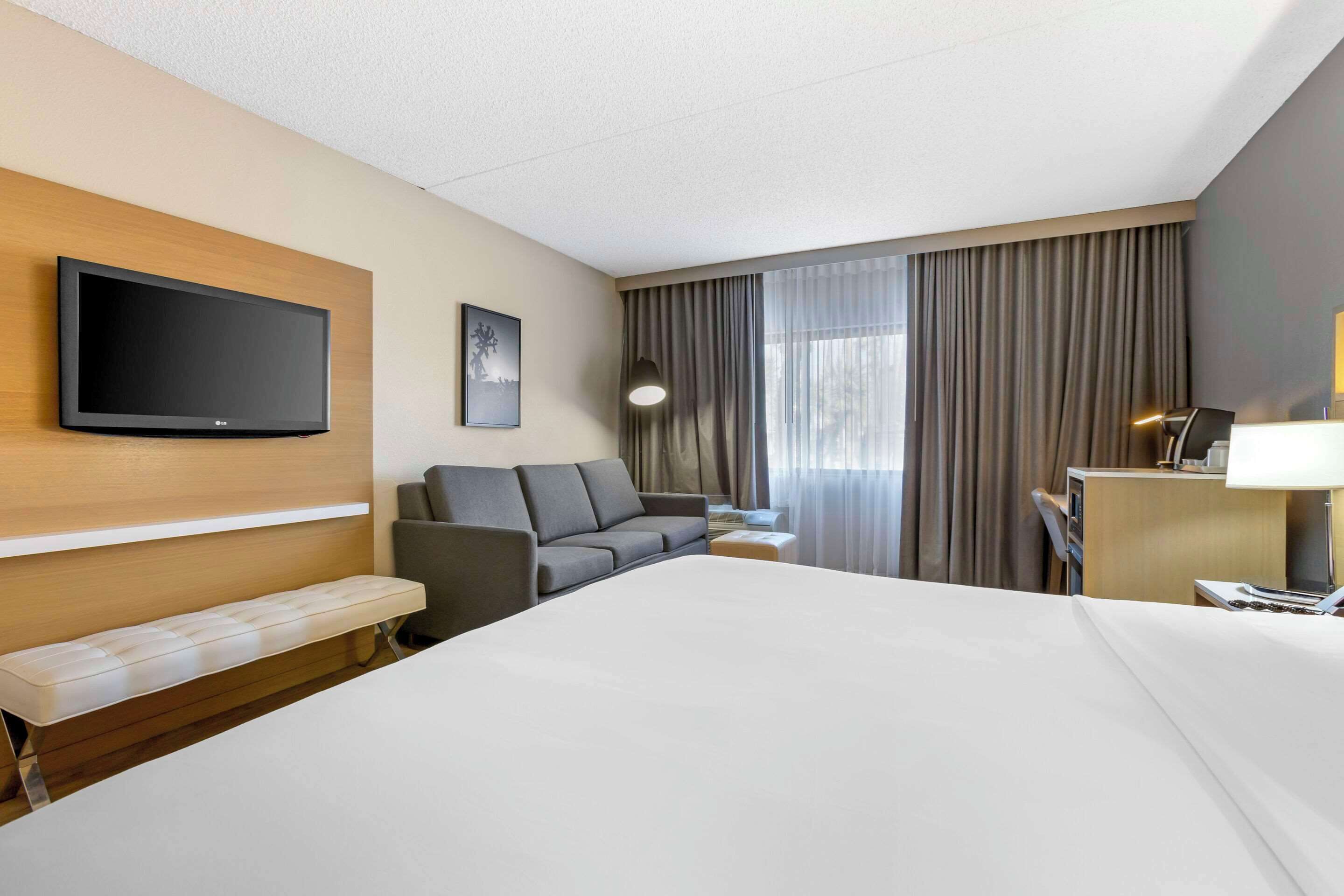 Quarto de hotel com uma cama grande, televisão de tela plana na parede, um sofá, uma escrivaninha com abajur e uma janela com cortinas.