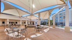 An outdoor patio area with tables and chairs, large umbrellas, and a pool in the background.