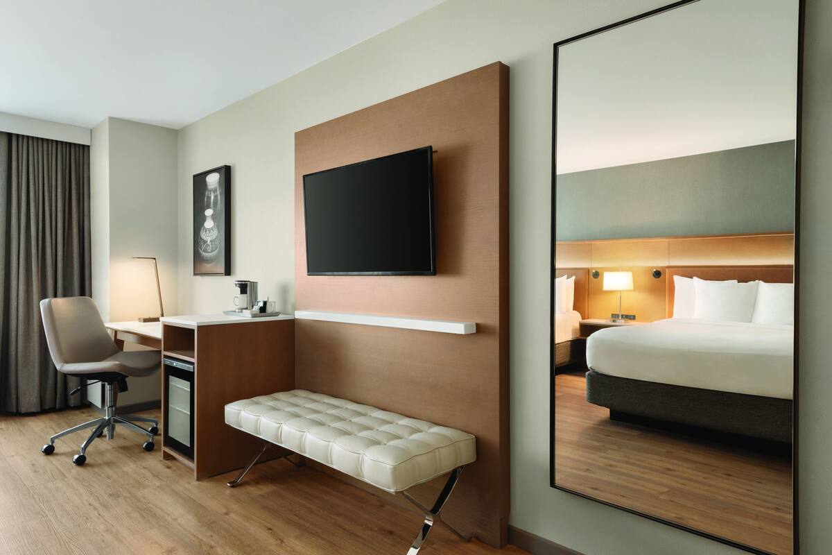 Habitación de hotel con una cama, un escritorio con silla, un banco, un televisor montado en la pared y una mesita de noche con lámpara.
