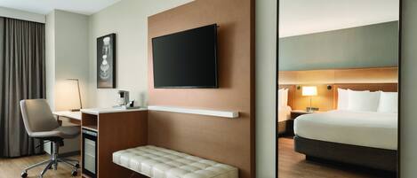 Habitación de hotel con una cama, un escritorio con silla, un banco, un televisor montado en la pared y una mesita de noche con lámpara.