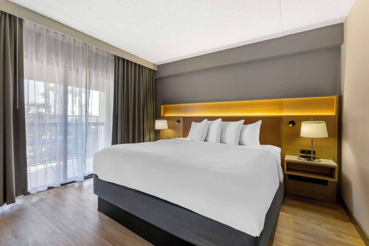 Una habitación de hotel moderna con una cama grande, una mesita de noche con lámpara y vistas a través de cortinas translúcidas.