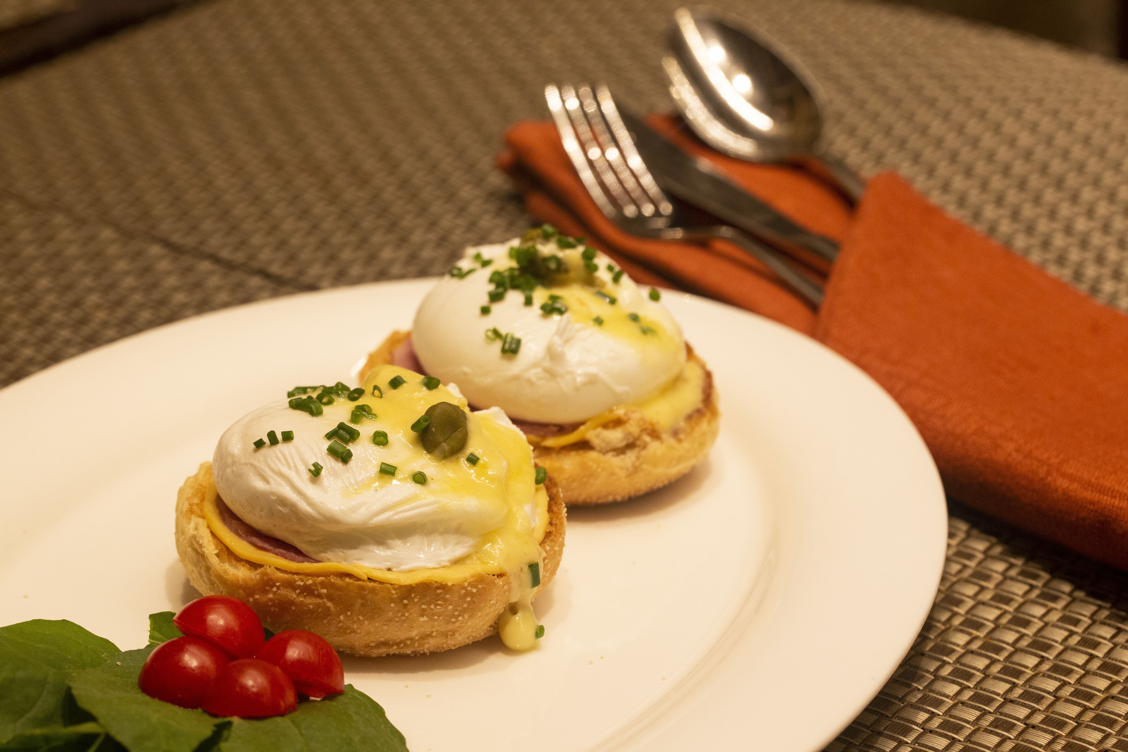 Dos sándwiches huevos benedictinos sobre un plato blanco, con tomates cherry y hojas verdes.