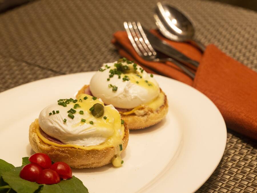 Dos sándwiches huevos benedictinos sobre un plato blanco, con tomates cherry y hojas verdes.