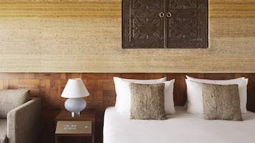 A hotel room with a wooden headboard, a bed with white linens and textured pillows, a nightstand with a lamp, and a chair.