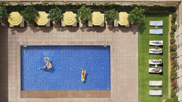 An aerial view of a swimming pool with people in the water, surrounded by lounge chairs and umbrellas.
