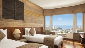 A hotel room with two beds, a sitting area with a table and chairs, a large window with a city and sea view, and a decorative wall panel. - President, Mumbai - IHCL SeleQtions (Mumbai)