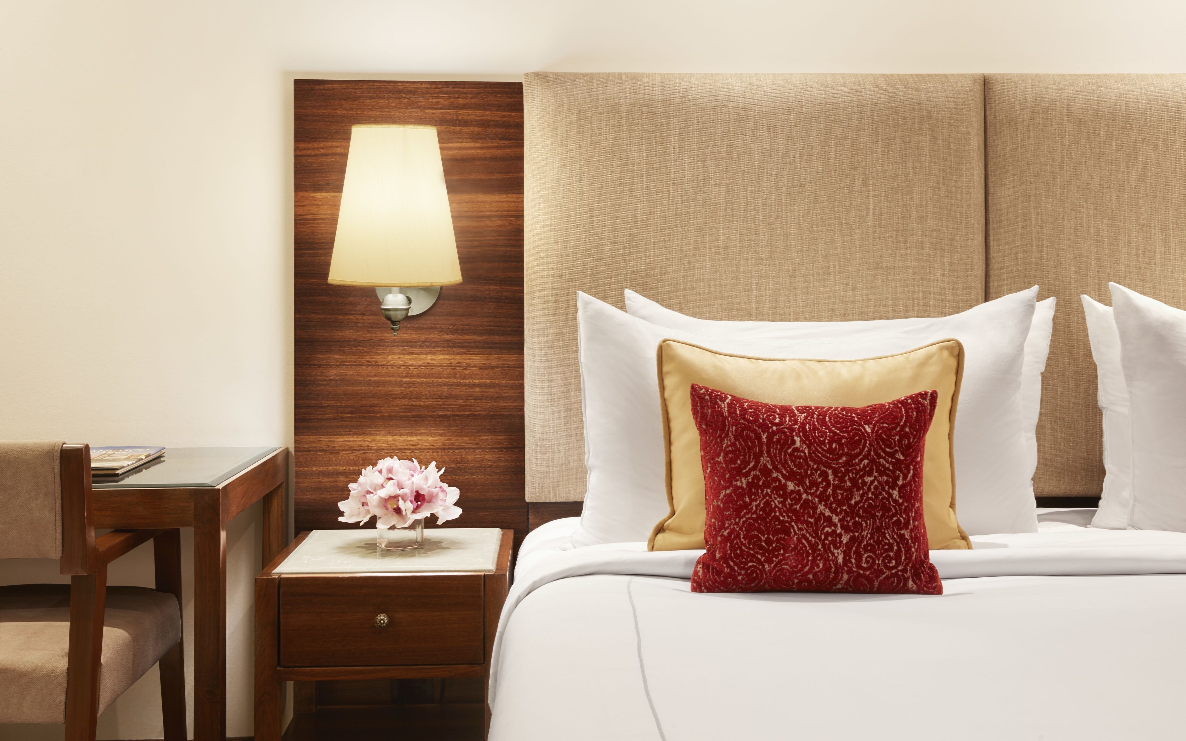 Een hotelkamer met een houten hoofdeinde, een nachtkastje met een lamp, een vaas met bloemen, en een bed met wit beddengoed en decoratieve kussens.