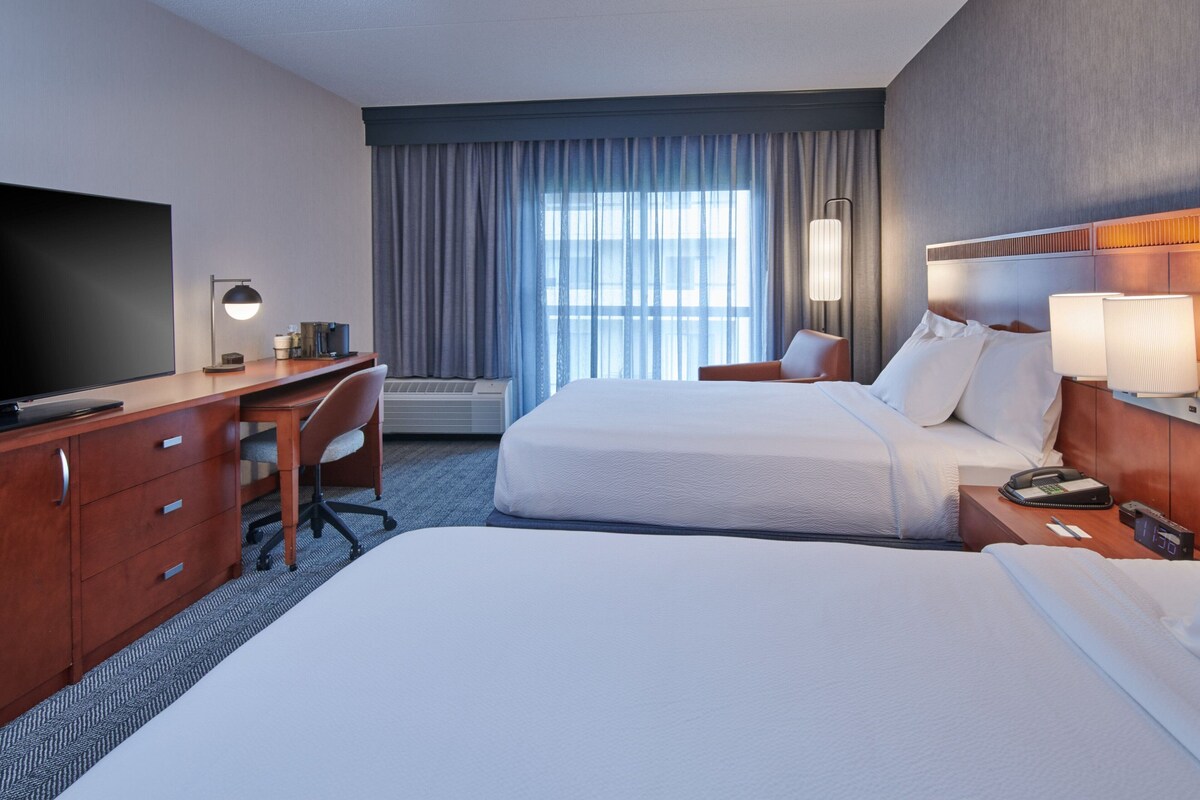 A hotel room with two beds, a desk, a TV, and a large window with curtains.