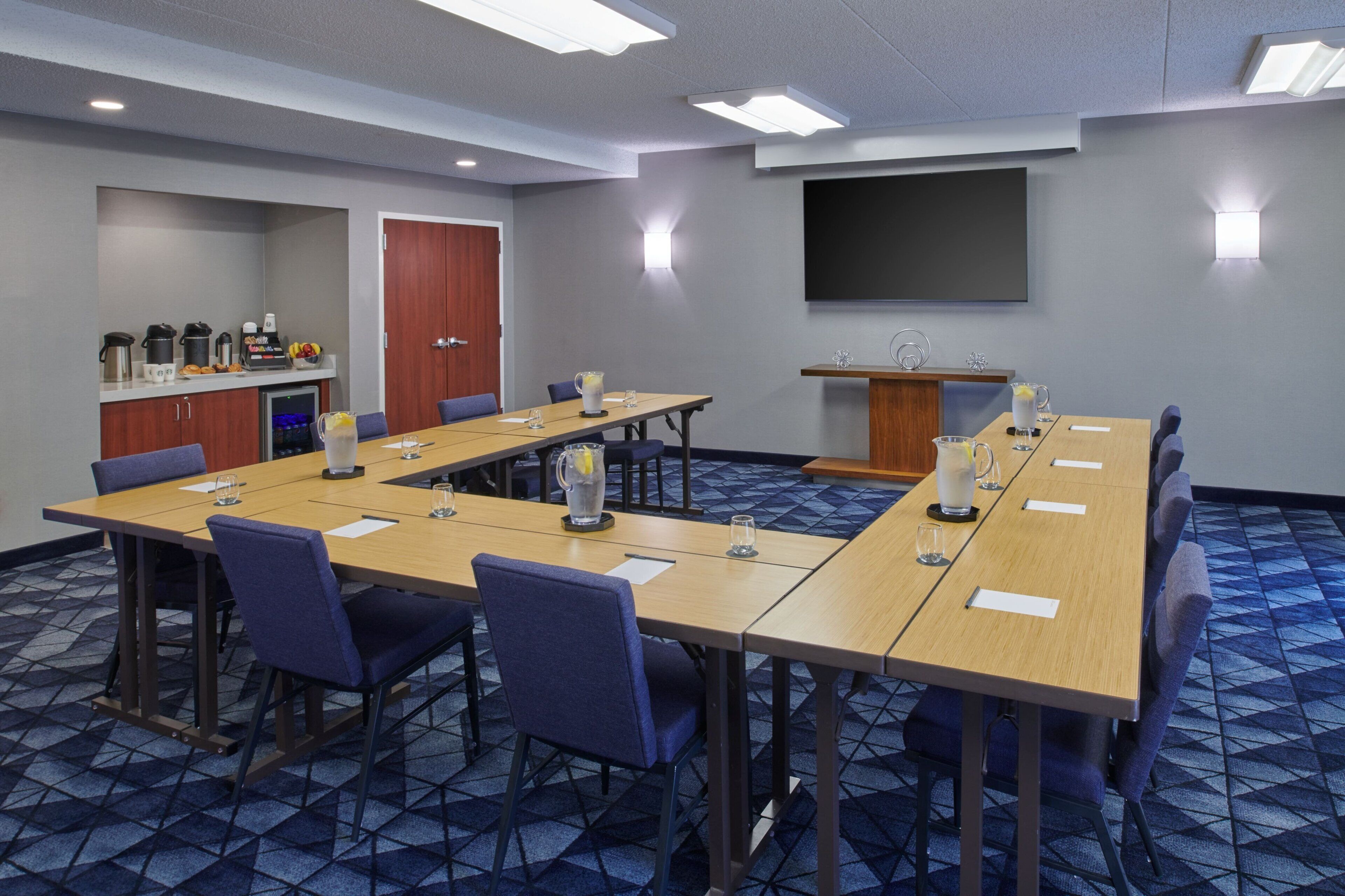 A conference room with a long table, chairs, a projector screen, and a kitchenette area.