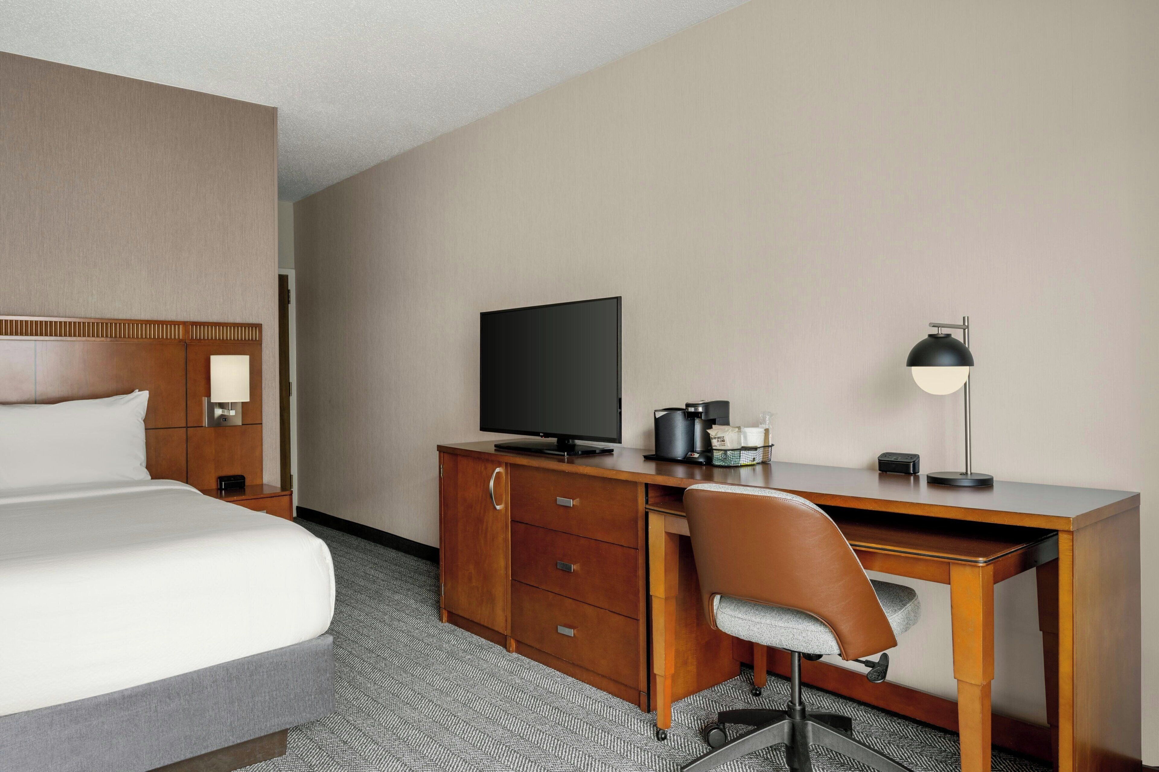 A hotel room with a bed, a desk with a television, a chair, and a lamp.