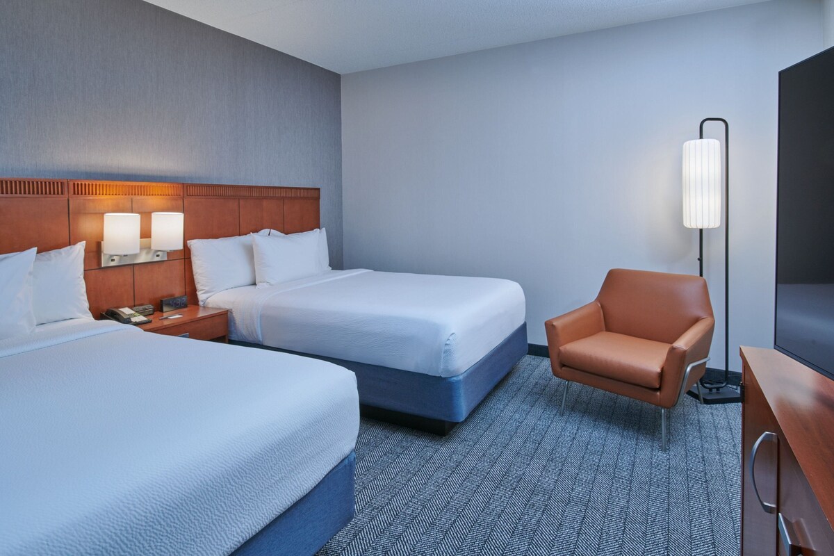 A hotel room with two beds, a nightstand, a chair, a desk, and a TV.