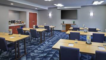 A conference room with multiple tables and chairs, a kitchenette, and a large screen on the wall.