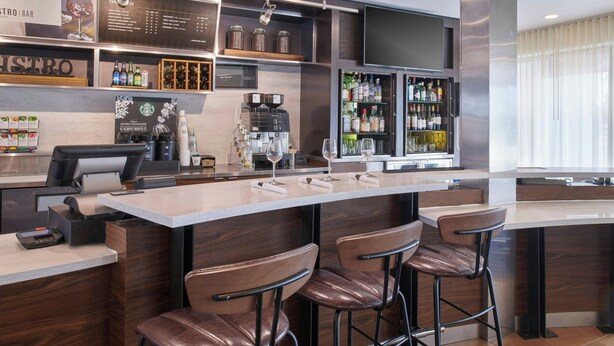 A modern bar area with a long counter, bar stools, a coffee machine, and a wine cooler. - Courtyard by Marriott Detroit Livonia (Livonia)