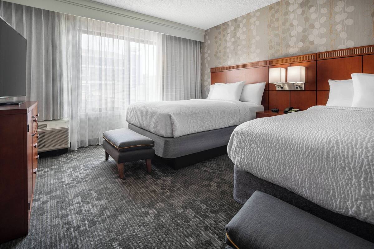 Habitación de hotel con dos camas, un escritorio, una silla y un ventanal con cortinas traslúcidas.