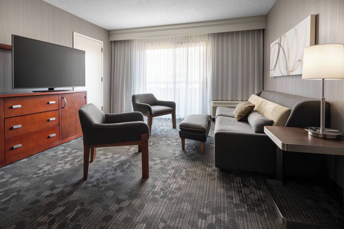 Una habitación de hotel con televisión de pantalla plana, un mueble de madera, dos sillones, un sofá, una mesita auxiliar y una lámpara.
