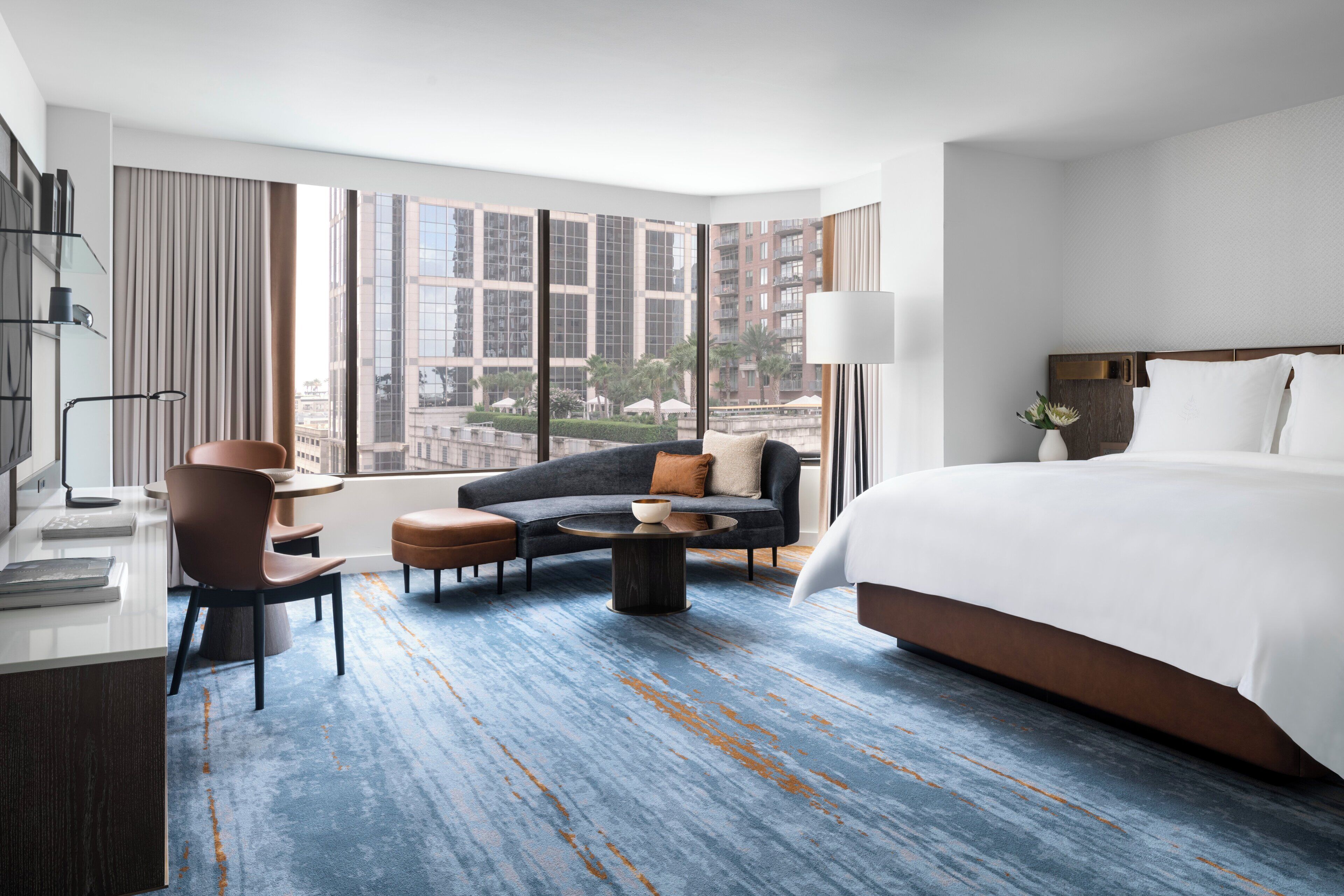 A hotel room with a large bed, a sofa, a coffee table, a desk with a lamp, and a view of the city through large windows.
