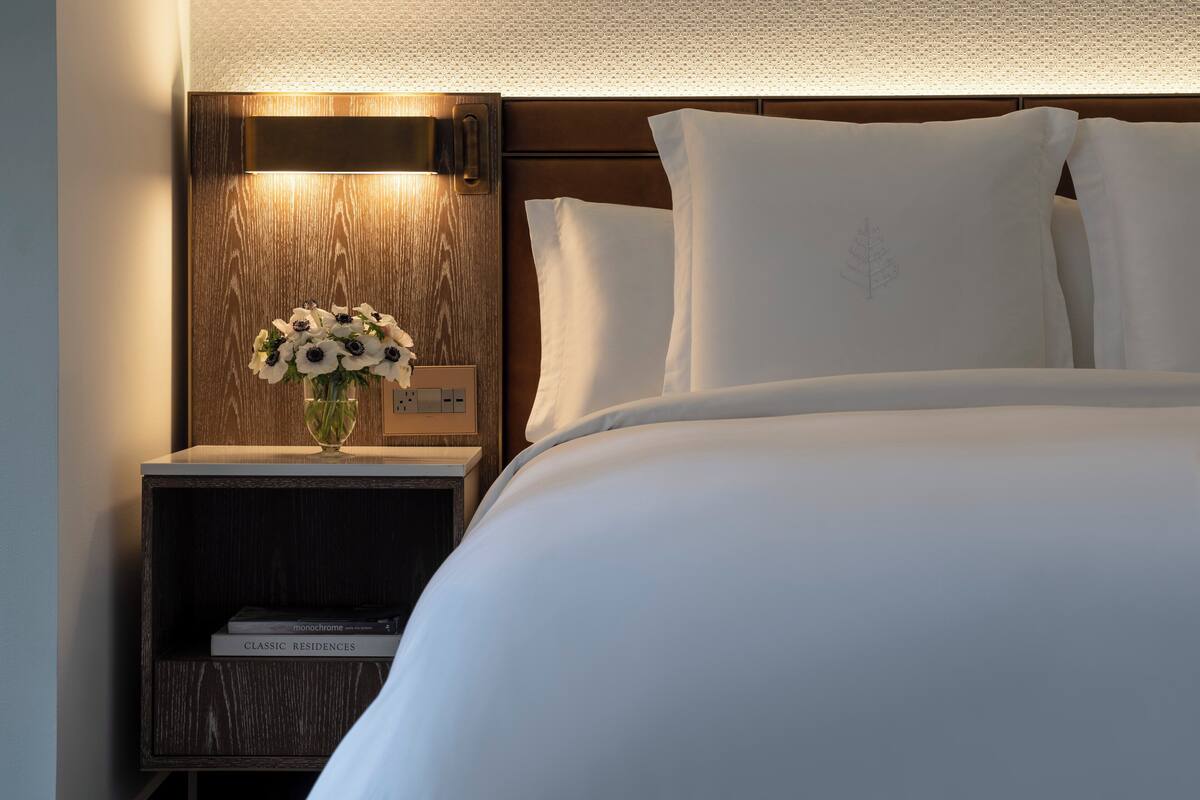 A neatly made bed with white linens, a wooden nightstand with a vase of flowers, and a wall-mounted light fixture.