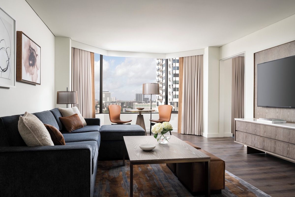 Une chambre d’hôtel moderne dotée d’une grande fenêtre, d’un canapé, d’une table basse, d’un téléviseur et d’un coin repas.