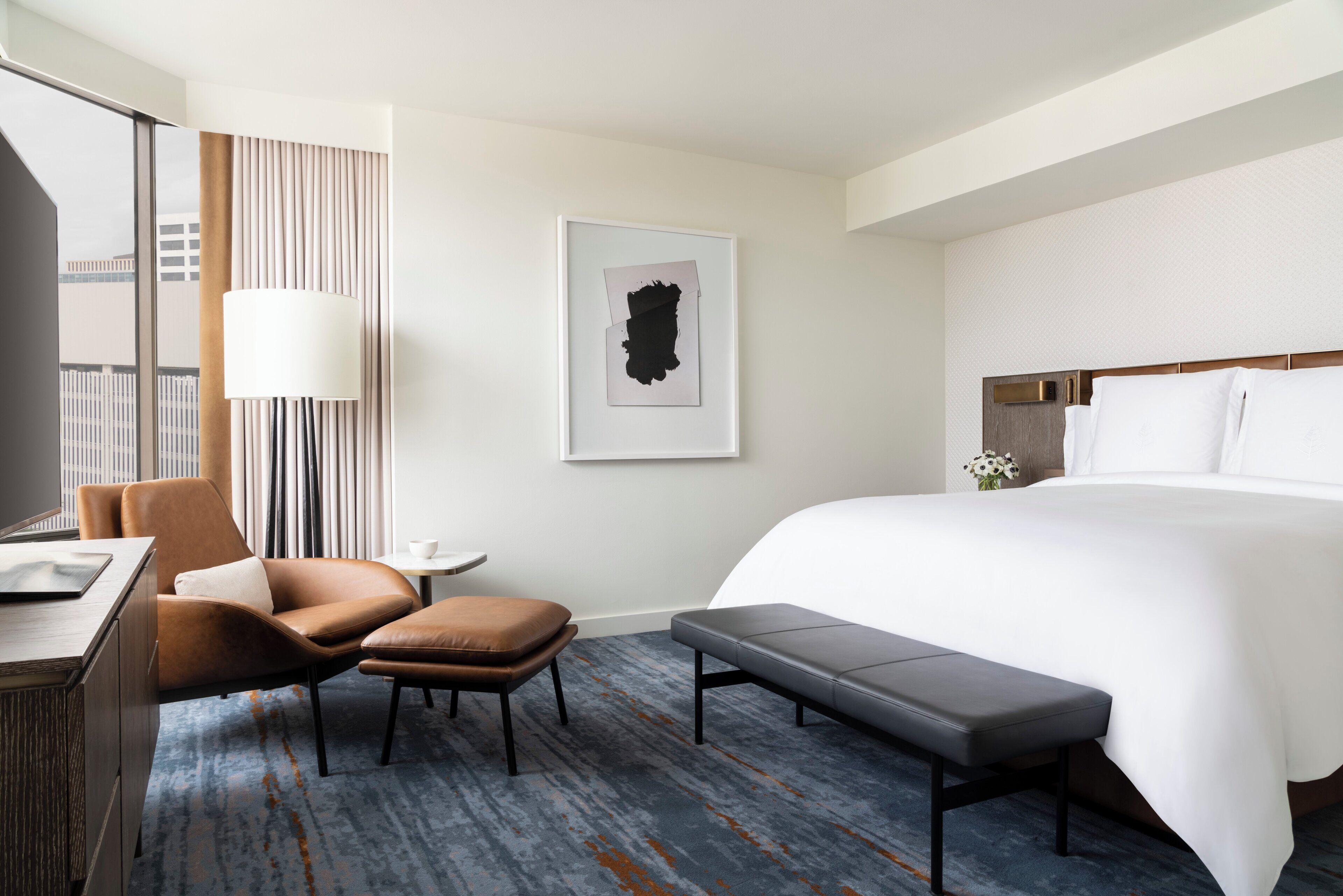 A modern hotel room with a large bed, a desk, a chair, and a bench.