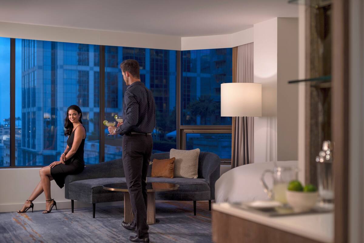 A modern hotel room with a large window offering a city view, a sofa, a coffee table, and a small side table with a lamp.