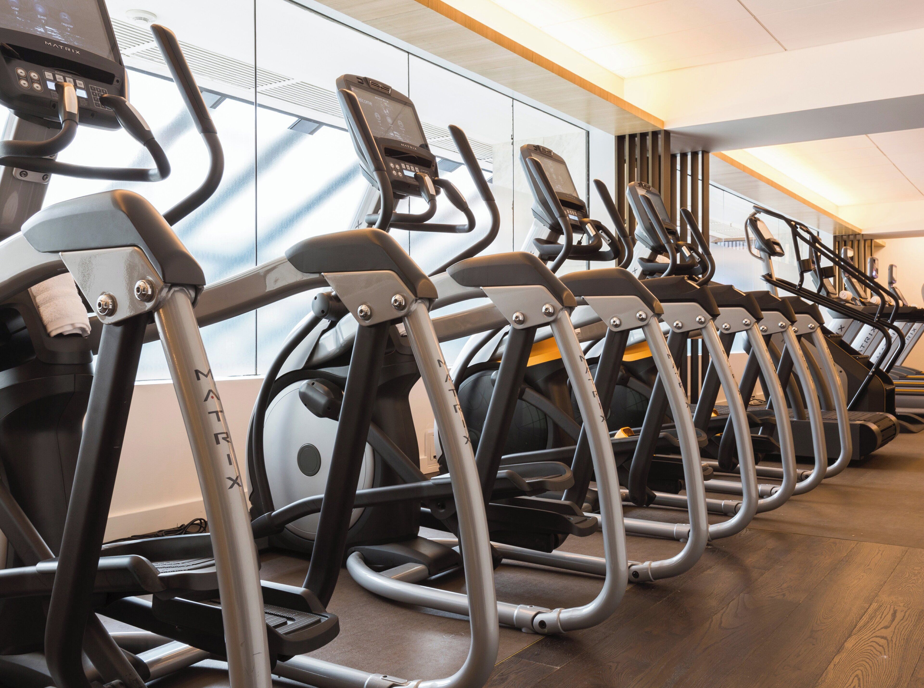 A row of stationary bikes in a gym with a mirrored wall and a wooden floor.